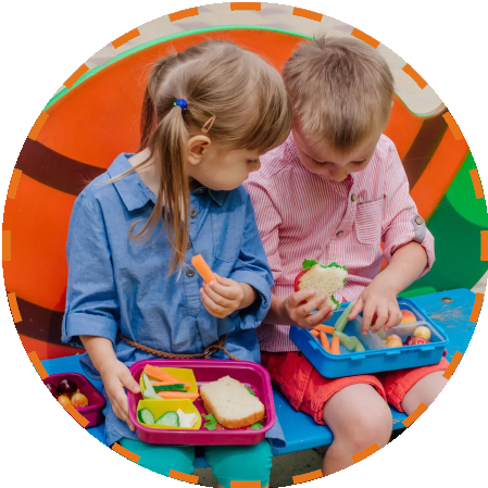 Group-1321316680.png (4) (4) Two children sitting on a bench eating food.
